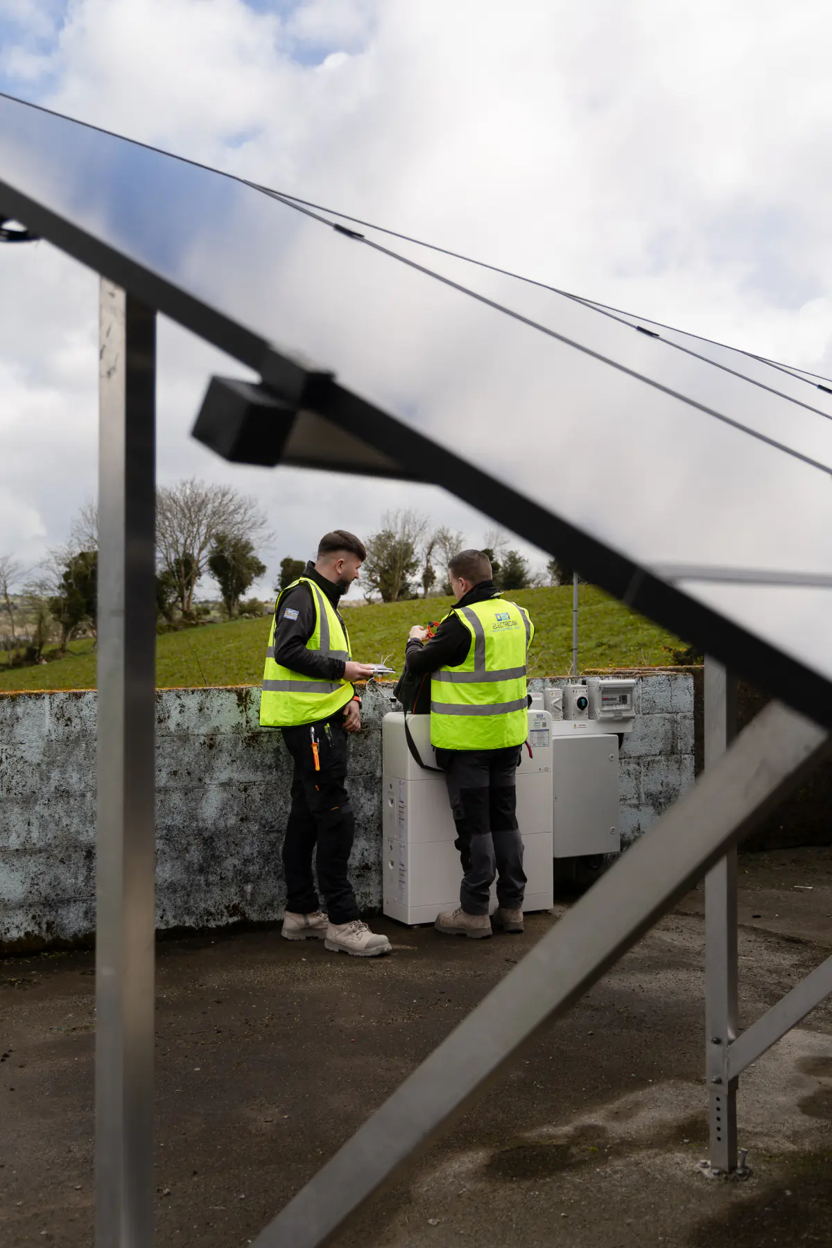 Two 021 Electrician team members working on a solar battery system below ground-mounted solar panels.